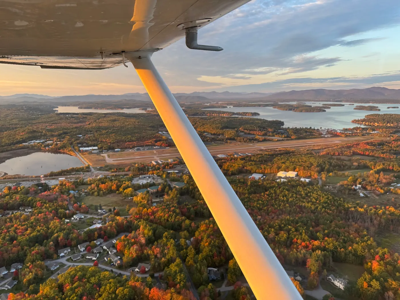 Aerial view from a small plane over Lake Winnipesaukee, New Hampshire, in fall, the wing strut in frame, foliage and a runway below
